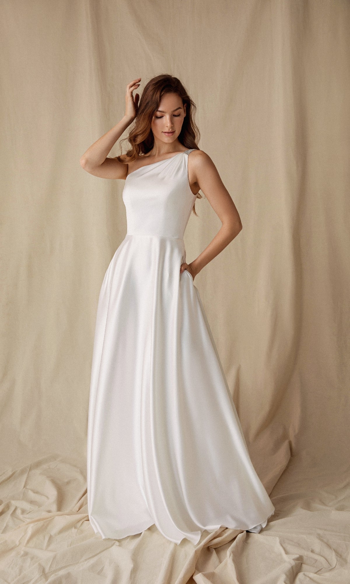 A woman in the Eureka 9025 One-Shoulder Long Satin Formal Dress poses against a beige fabric backdrop, one hand in her pocket and the other touching her hair.